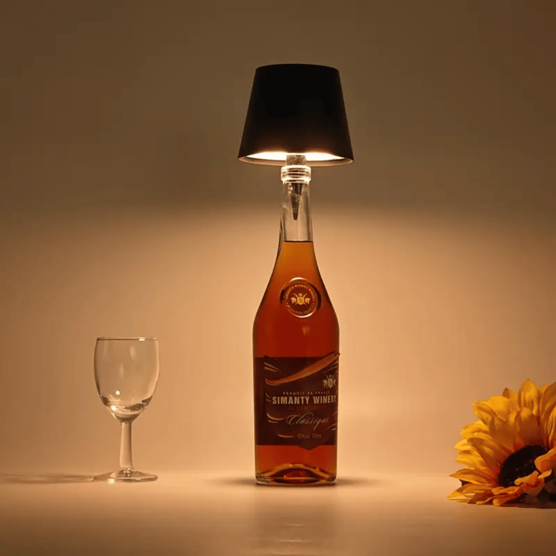 An elegant lamp made from a bottle with a black lampshade, illuminated to cast a warm light, placed on a surface next to a glass and a sunflower.