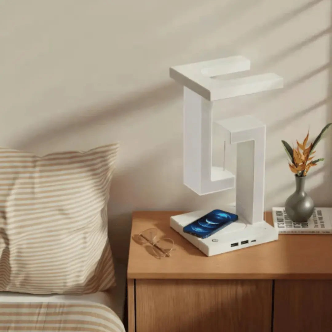 A white LED desk lamp with a built-in wireless charger on a wooden table next to a book and a smartphone.