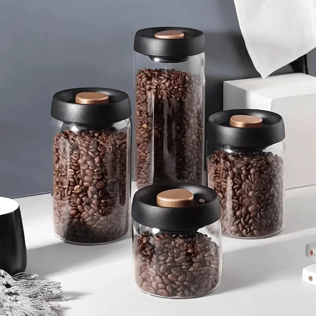 A set of press-seal glass canisters of various sizes with matte black lids featuring a luxurious gold accent, filled with what appears to be coffee beans.