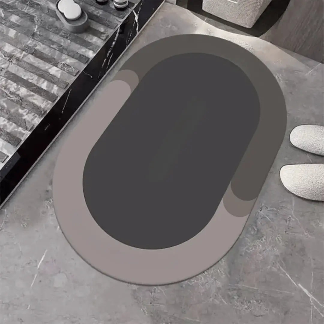 A gray oval-shaped bathroom mat placed on a tiled floor, with a pair of slippers at the top right corner.