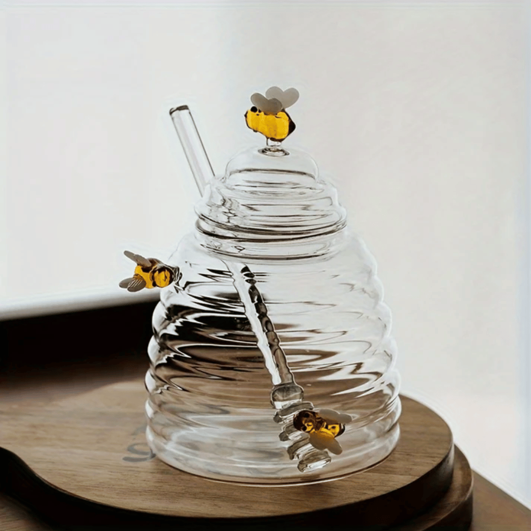 A person is pouring liquid from a clear glass jar with a honeycomb pattern into another container, using a metal honey dipper.