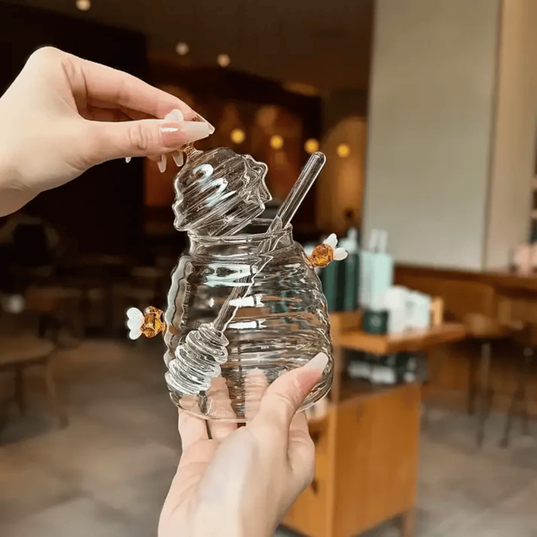 A person is pouring liquid from a clear glass jar with a honeycomb pattern into another container, using a metal honey dipper.