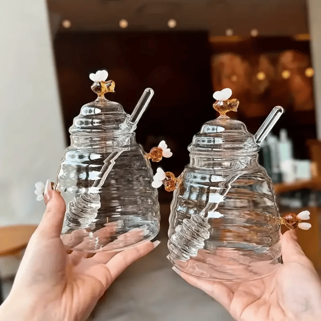 A person is pouring liquid from a clear glass jar with a honeycomb pattern into another container, using a metal honey dipper.