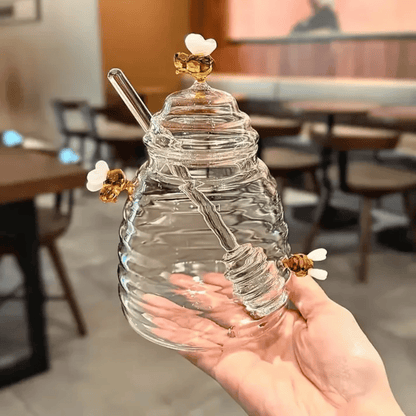 A person is pouring liquid from a clear glass jar with a honeycomb pattern into another container, using a metal honey dipper.