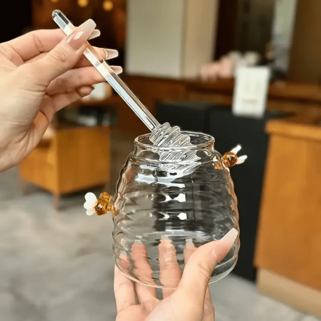 A person is pouring liquid from a clear glass jar with a honeycomb pattern into another container, using a metal honey dipper.