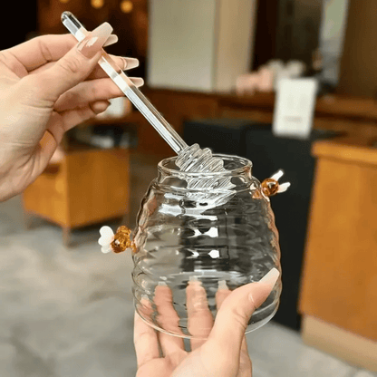 A person is pouring liquid from a clear glass jar with a honeycomb pattern into another container, using a metal honey dipper.