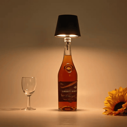 An elegant lamp made from a bottle with a black lampshade, illuminated to cast a warm light, placed on a surface next to a glass and a sunflower.