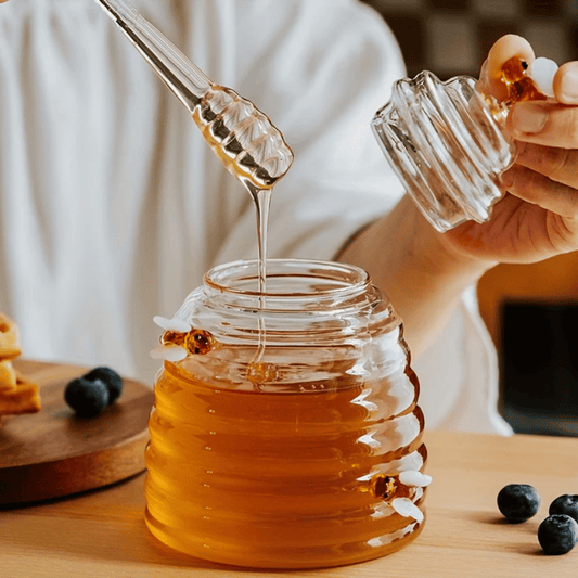 A person is pouring liquid from a clear glass jar with a honeycomb pattern into another container, using a metal honey dipper.