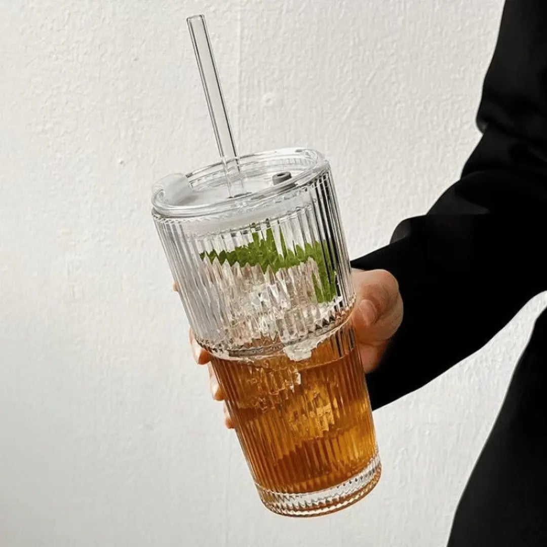 Hand holding a clear ribbed tumbler with a drink and straw against a light background