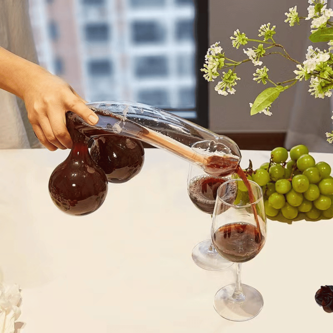 A clear glass decanter in a humorous design resembling a figure, with two filled wine glasses beside it, placed on a table with a window in the background.