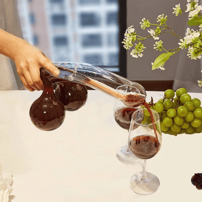 A clear glass decanter in a humorous design resembling a figure, with two filled wine glasses beside it, placed on a table with a window in the background.