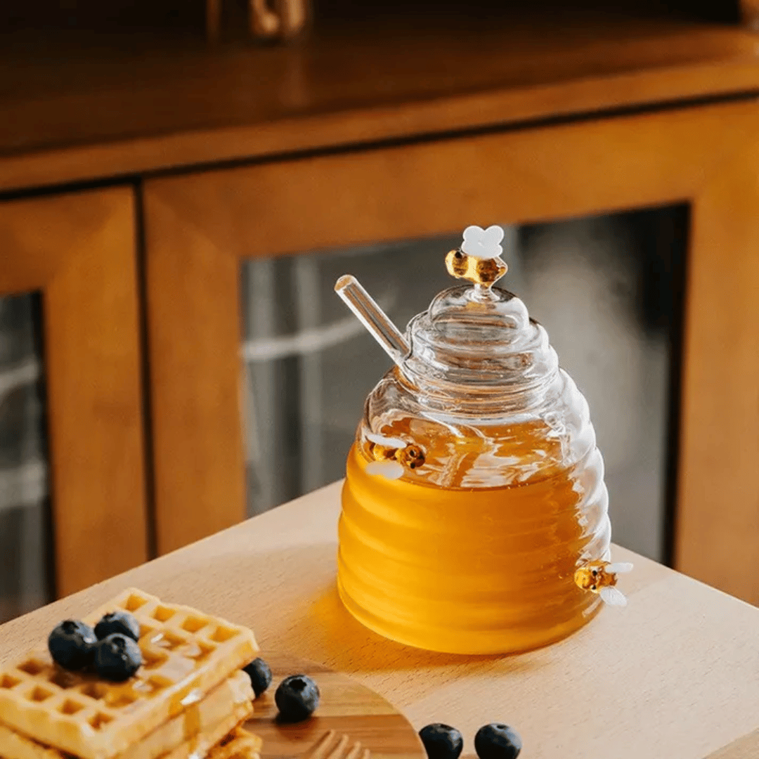 A person is pouring liquid from a clear glass jar with a honeycomb pattern into another container, using a metal honey dipper.