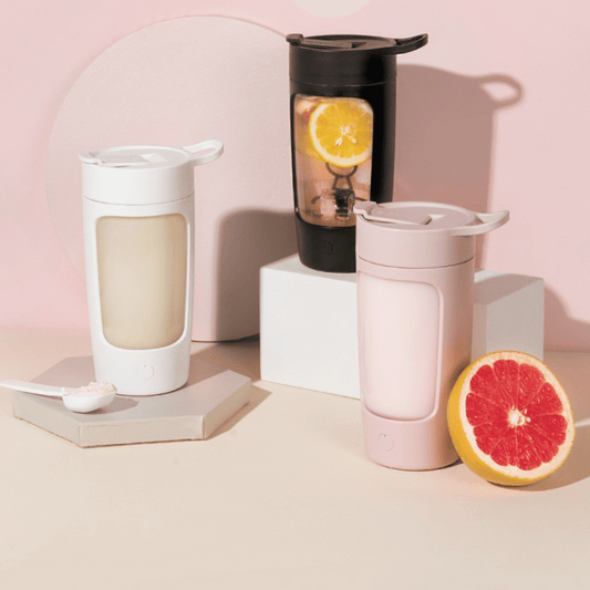 Three portable mixers in white, black, and pink colors, displayed on a neutral background with a cut grapefruit and a lemon slice.
