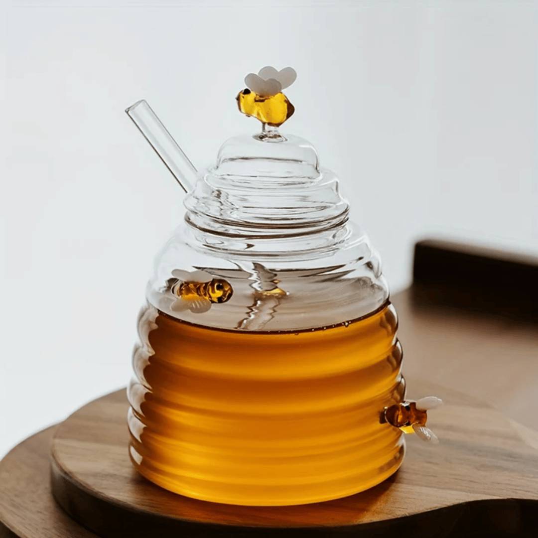 A person is pouring liquid from a clear glass jar with a honeycomb pattern into another container, using a metal honey dipper.