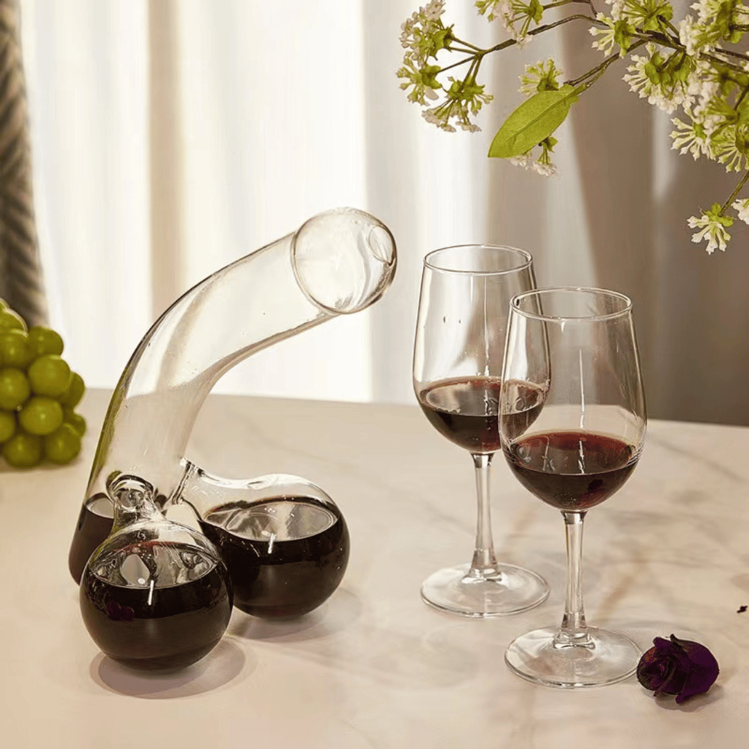 A clear glass decanter in a humorous design resembling a figure, with two filled wine glasses beside it, placed on a table with a window in the background.