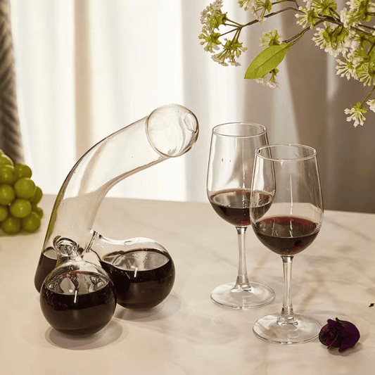 A clear glass decanter in a humorous design resembling a figure, with two filled wine glasses beside it, placed on a table with a window in the background.