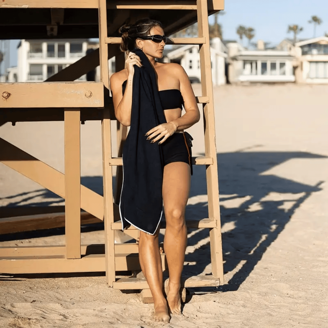 Woman in black swimsuit and towel standing on a beach.