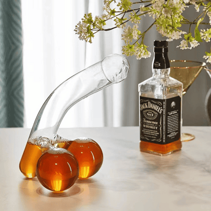 A clear glass decanter in a humorous design resembling a figure, with two filled wine glasses beside it, placed on a table with a window in the background.