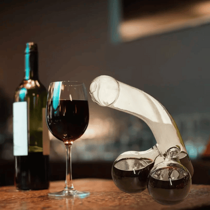 A clear glass decanter in a humorous design resembling a figure, with two filled wine glasses beside it, placed on a table with a window in the background.