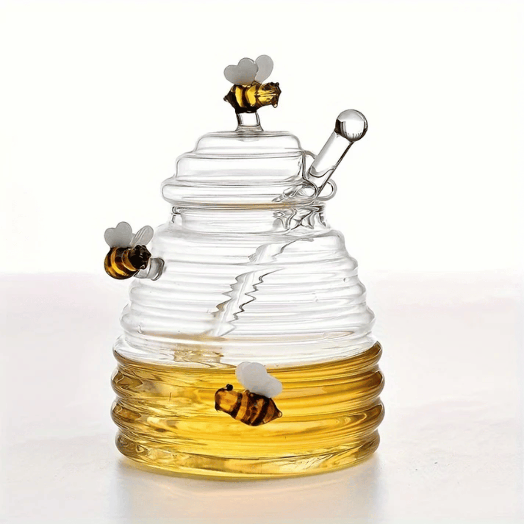 A person is pouring liquid from a clear glass jar with a honeycomb pattern into another container, using a metal honey dipper.