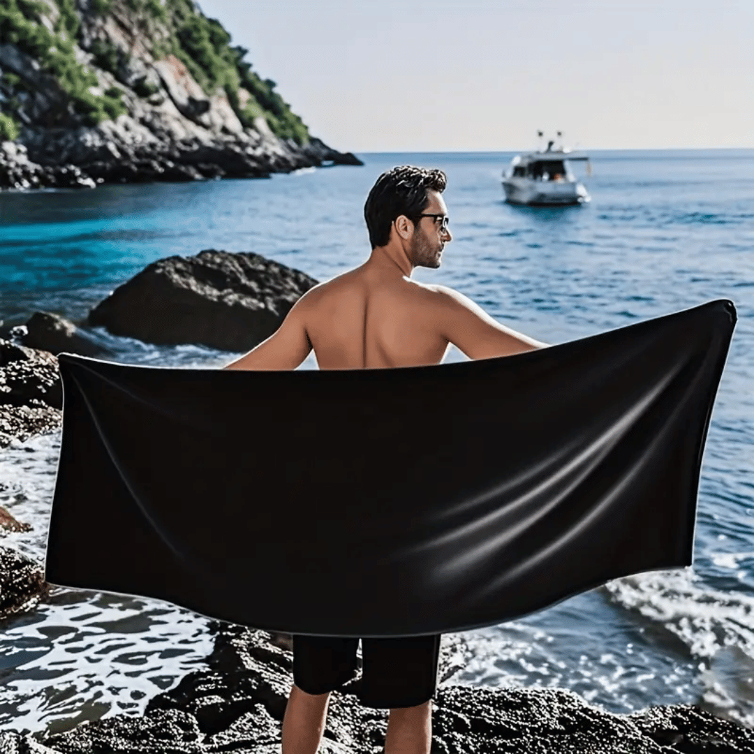 Man on a rocky beach holding a black towel with a boat in the background.