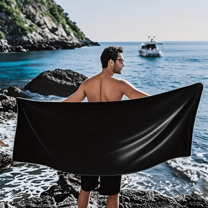 Man on a rocky beach holding a black towel with a boat in the background.