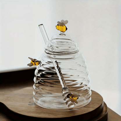 A person is pouring liquid from a clear glass jar with a honeycomb pattern into another container, using a metal honey dipper.