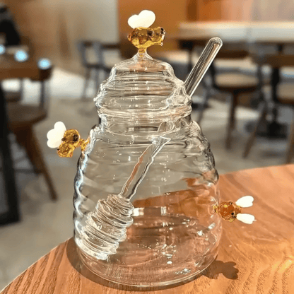 A person is pouring liquid from a clear glass jar with a honeycomb pattern into another container, using a metal honey dipper.